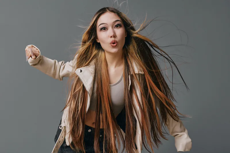 Young woman with long hair posing in studio wearing casual jacket and top, motion captured with hair flying and dynamic expression on neutral gray background.