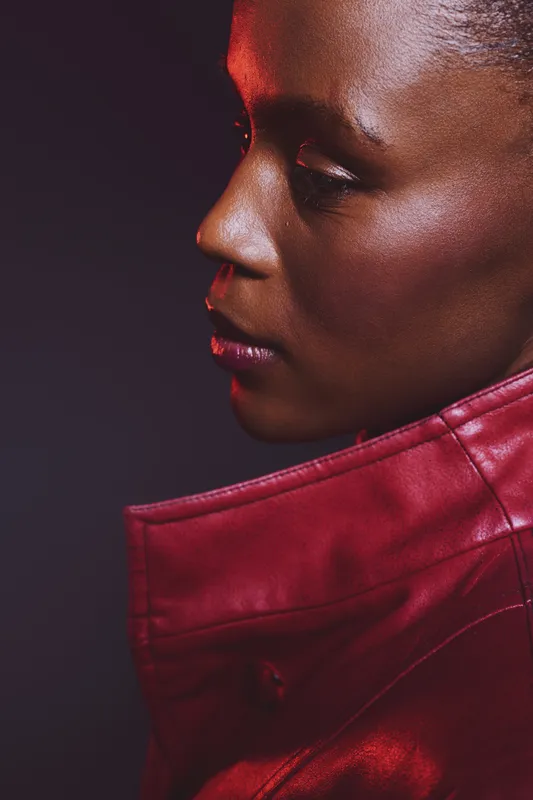 Close-up profile of a dark skinned African woman wearing a red leather jacket. The studio portrait captures her smooth skin and thoughtful expression on a dark background.