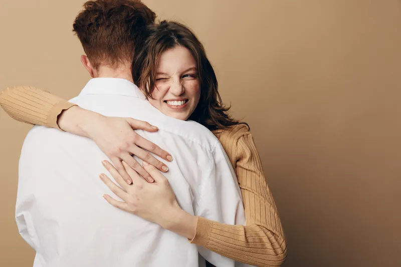 Happy woman in beige sweater hugs man in white shirt, smiling with eyes closed. Intimate moment of affection and warmth against plain beige background, concept of love and connection.