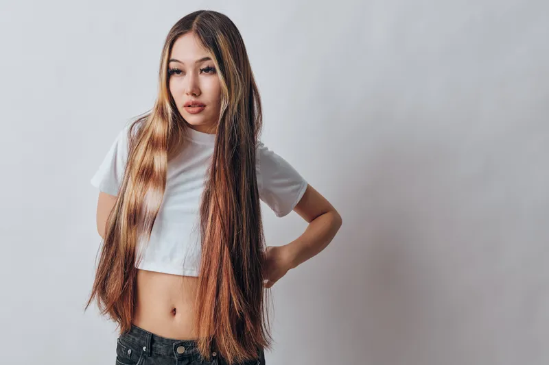 Young woman with long hair wearing white crop top and black pants standing with hand on hip looking thoughtful on plain background.