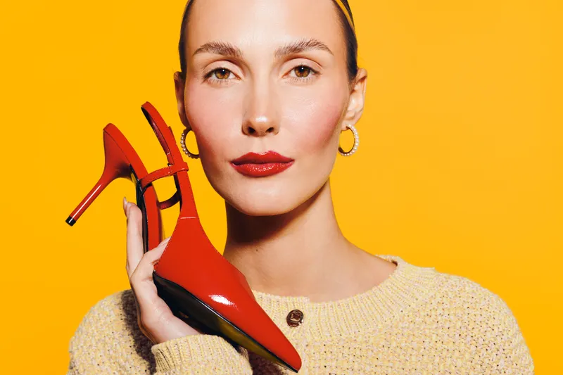Close-up of confident woman holding red high heel shoe near face on yellow background. Concept of fashion, style, beauty, and elegance with vibrant colors and minimalistic composition.