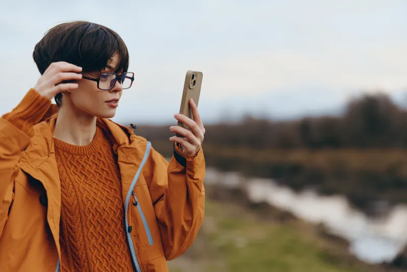 Woman outdoors near river holding smartphone for selfie, wearing glasses and an orange jacket, close portrait in natural light, calm landscape, lifestyle and communication moment