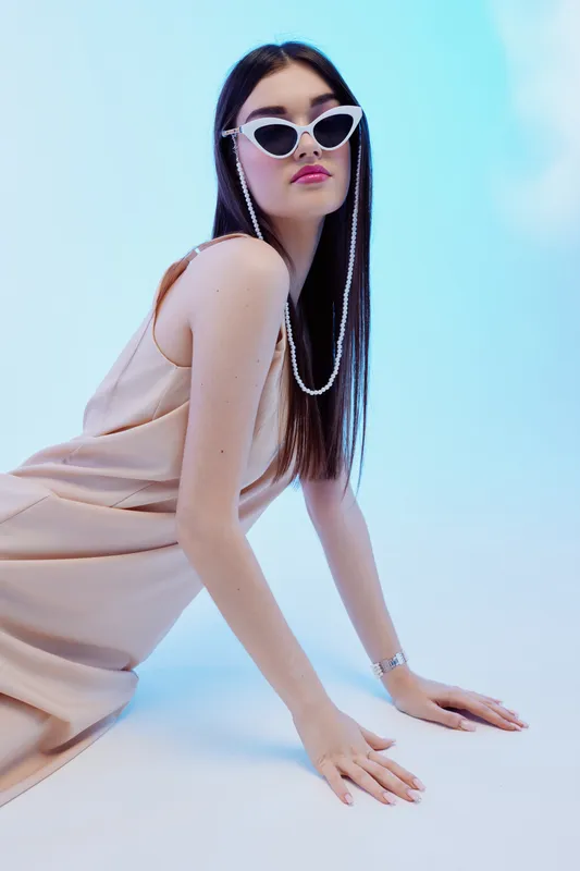 Fashionable woman with long straight hair wearing white sunglasses and a beige dress poses on a light blue background. Concept of beauty, fashion, and modern style.