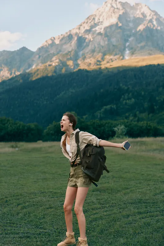 Young woman with backpack laughs joyfully while standing on a grassy field with mountains in the background during a sunny day outdoors.
