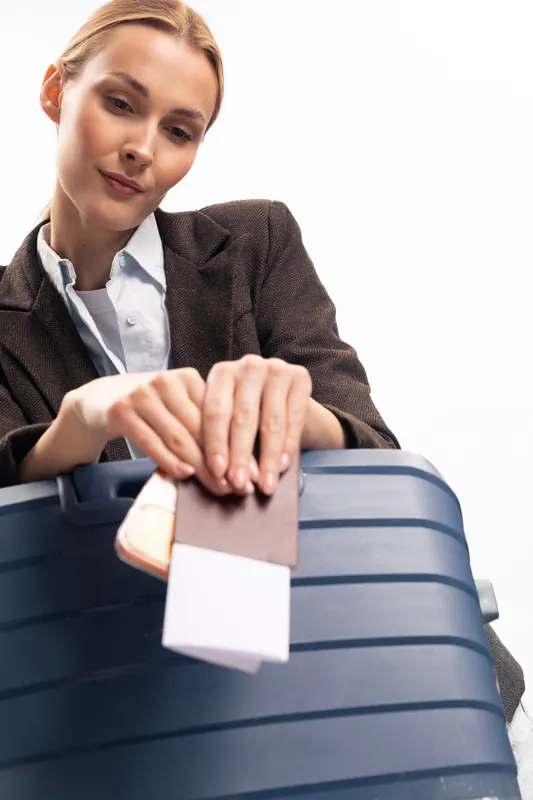 Young woman in formal jacket holds passport and boarding pass while packing blue suitcase. Travel concept for business trip, tourism, and vacation preparation on white background.