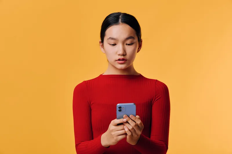 Young woman in red sweater holding smartphone and texting message against orange background. Portrait of female using mobile phone for communication with focused expression indoors.
