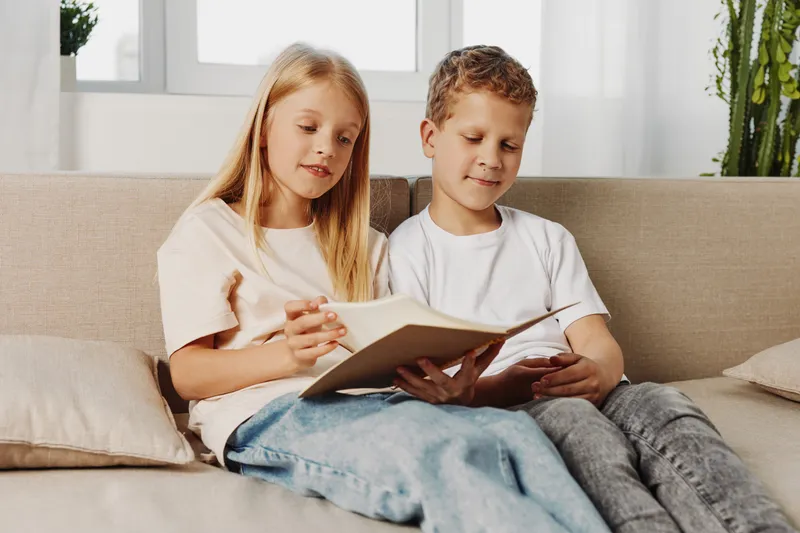 Two children sitting on a sofa reading a book together in a bright living room. The girl and boy share a calm moment of learning and curiosity, concept of childhood education and bonding.