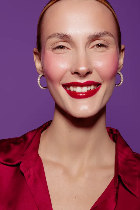 Smiling woman with red lipstick and hoop earrings wears red blouse against purple background. Close-up portrait with bright makeup and joyful expression. Concept of beauty and fashion.