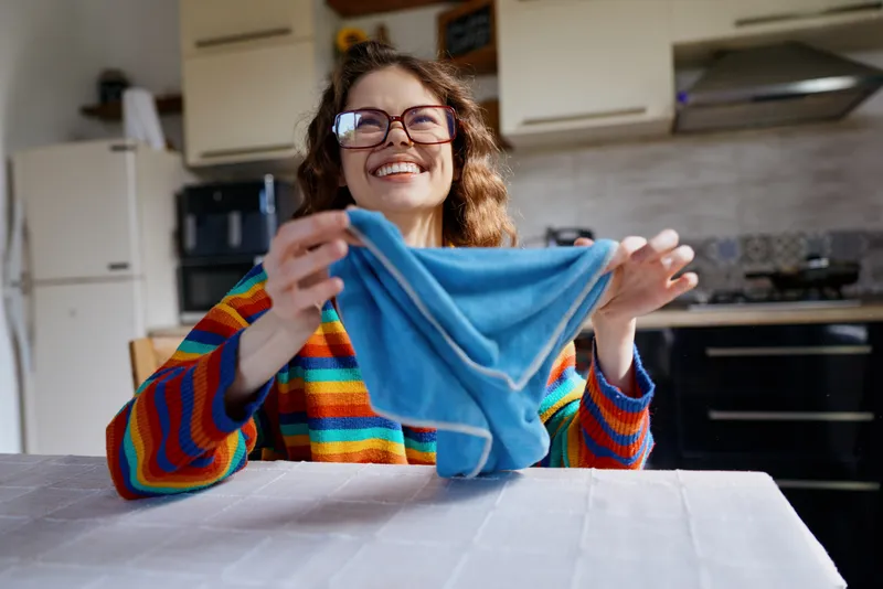 Woman with rainbow sweater sits at kitchen table, smiling while holding a blue cloth, cozy home moment, bright natural light, casual daily task, warm atmosphere.