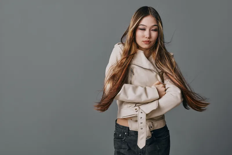 Young woman with long hair wearing light jacket and black jeans posing with crossed arms and closed eyes on gray background in studio portrait.