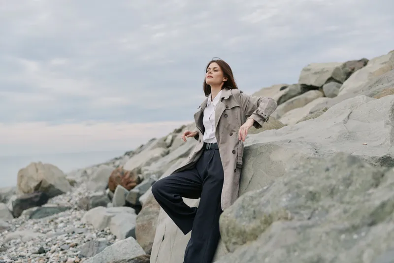 Young woman in stylish coat and black pants leans on large rocks by the sea shore. Overcast sky and rocky beach create a calm, contemplative mood in natural outdoor setting.