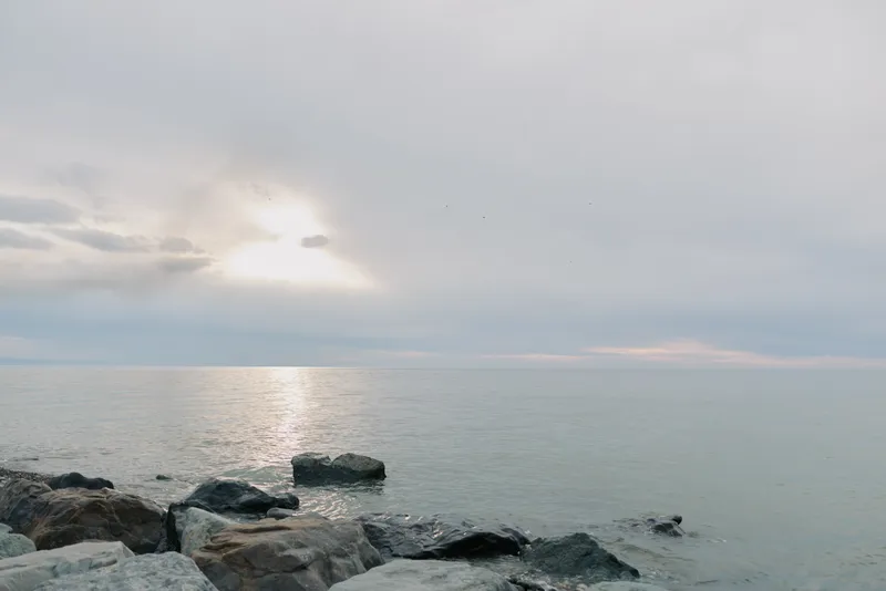 Calm sea with gentle waves and rocks in foreground under cloudy sky at sunrise. Peaceful coastal landscape with soft natural light reflecting on water surface.