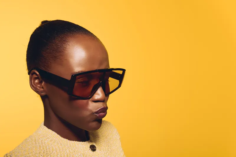 Portrait of a confident dark skinned African woman wearing large black sunglasses and a yellow sweater, posing against a bright yellow background. Concept of fashion and style.