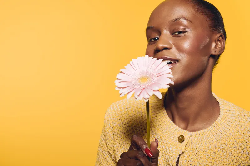 Smiling dark skinned African woman holds pink flower near face on yellow background. Concept of beauty, natural skin, skincare, happiness, and floral wellness in studio lighting.