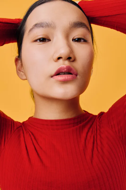 Young woman with clear skin and natural makeup poses with arms raised wearing a red shirt against yellow background. Minimalist style portrait with confident expression and soft lighting.