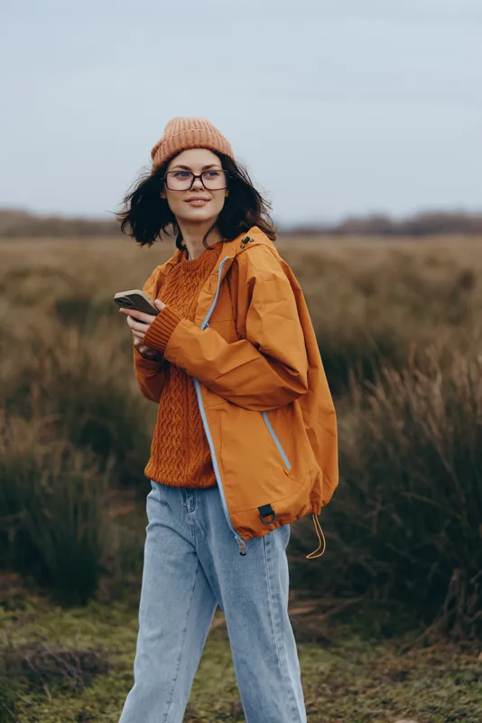 Smiling woman outdoors in a rural field, wearing beanie, glasses, orange sweater and jacket, holding phone while walking, casual lifestyle, overcast light, natural grass background