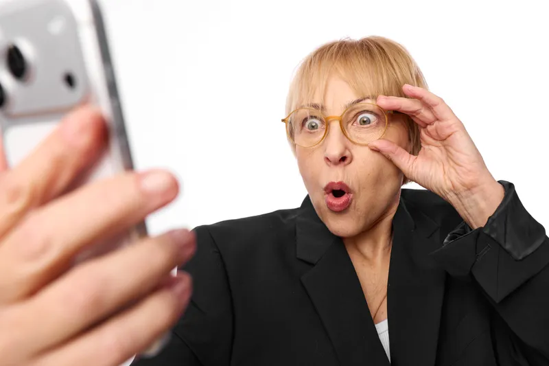 Elderly woman with glasses holds a smartphone, looking shocked and surprised, hand near her face, wearing a business jacket on a clean white background, communication moment.