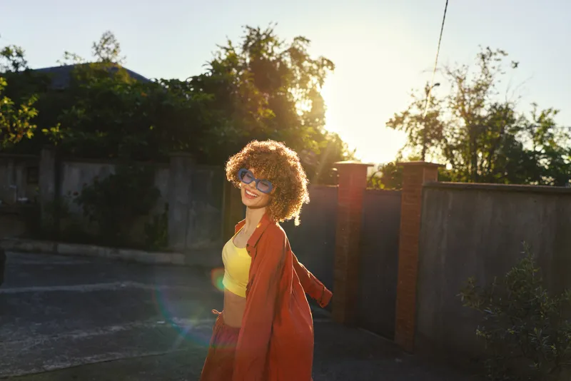 Cheerful woman with curly hair wearing a yellow top and red coat walks outdoors in warm sunlight. A lifestyle portrait focused on confidence, outdoor living, and self expression and joy