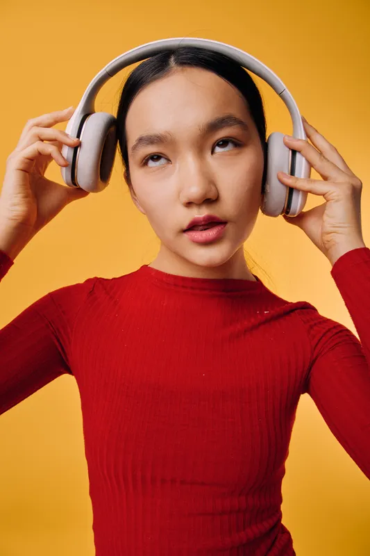 Young woman wearing headphones with hands adjusting them, standing against bright yellow background. Portrait of girl with black hair in red top listening to music or audio with focused expression.