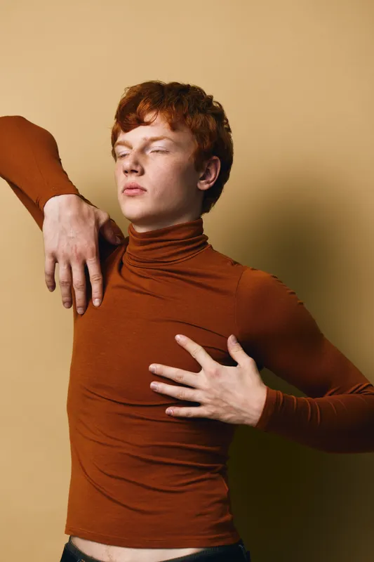 Young man with red hair wearing brown turtleneck posing with closed eyes and hands on chest and shoulder against beige background in studio portrait.