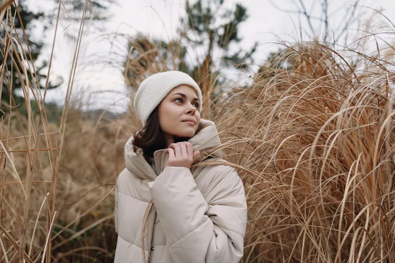 Winter woman in a creamy coat stands among tall grasses, gazing upward with a serene, cozy expression in a calm outdoor portrait among natural reeds.