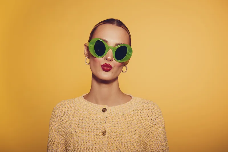 Stylish woman wearing green cucumber eye masks and red lipstick poses against yellow background. Concept for skincare, beauty, wellness, and self-care with fresh natural ingredients.