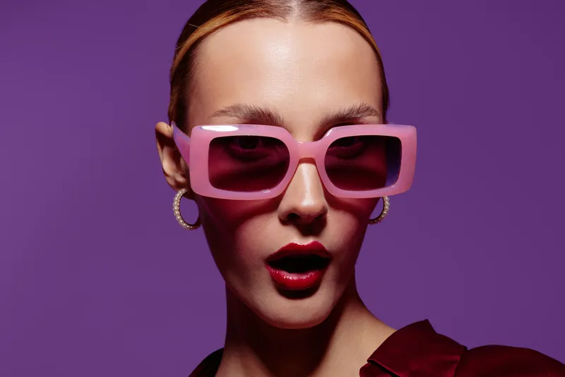 Stylish woman with pink oversized sunglasses and red lipstick poses on purple background. Fashion portrait with bold makeup and trendy accessories. Concept of modern style and beauty.