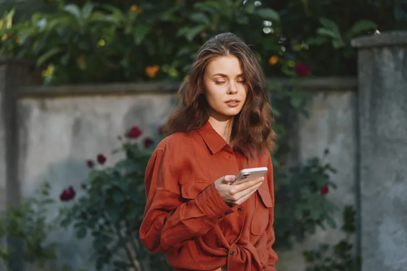 A woman in an orange shirt checks her phone in a sunlit garden, capturing a casual, everyday lifestyle moment outdoors, with relaxed posture and natural light