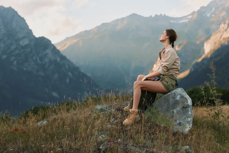 Young woman sits on a rock in a mountain meadow, gazing up with a calm, reflective mood, as soft natural light highlights her hiking outfit and the expansive alpine landscape.