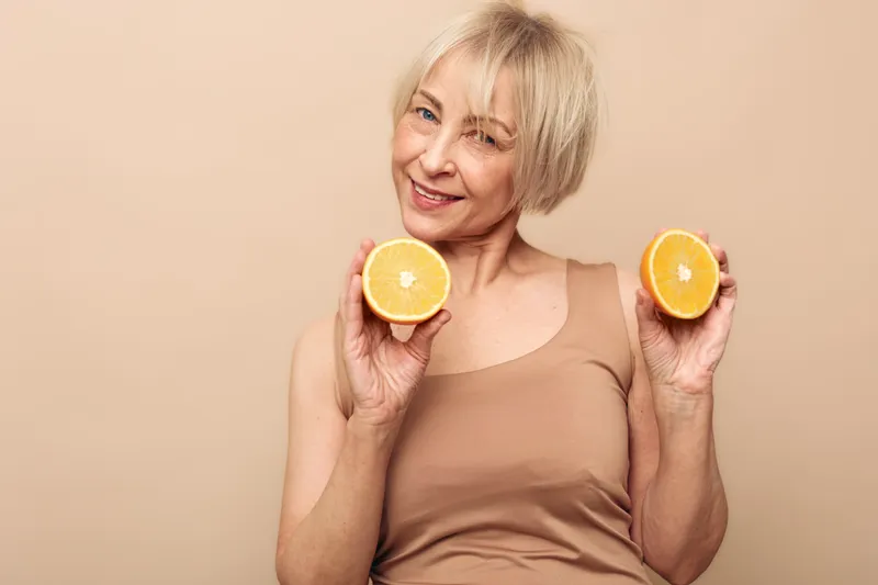 Smiling mature woman with short blonde hair holds two halves of fresh orange fruit near face against beige background. Concept of healthy lifestyle, vitamin C, wellness, and natural skincare.