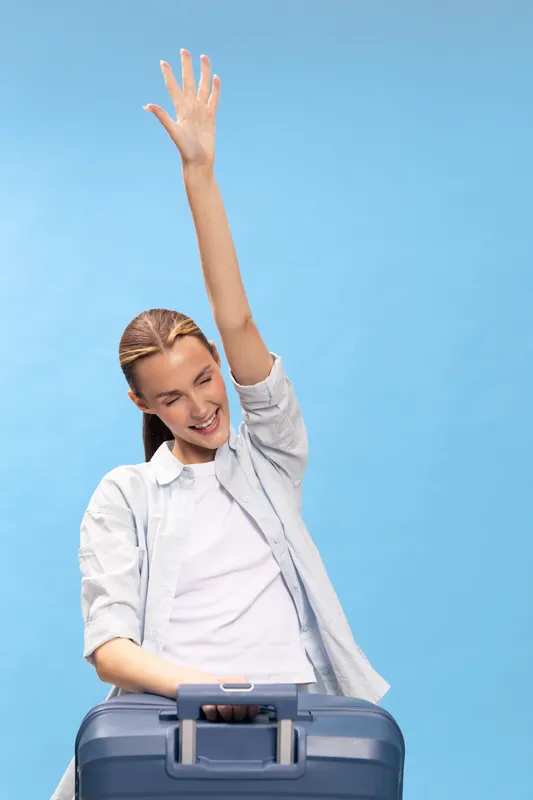 Happy young woman with suitcase raising hand smiling, dressed in casual clothes, isolated on blue background. Concept for travel, vacation, joy, freedom, and adventure.