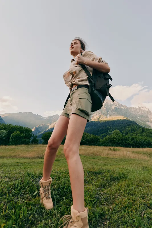Hiking woman with backpack explores a meadow near mountains, wearing shorts and boots, enjoying outdoor adventure, fresh air, and a scenic landscape on a sunny day