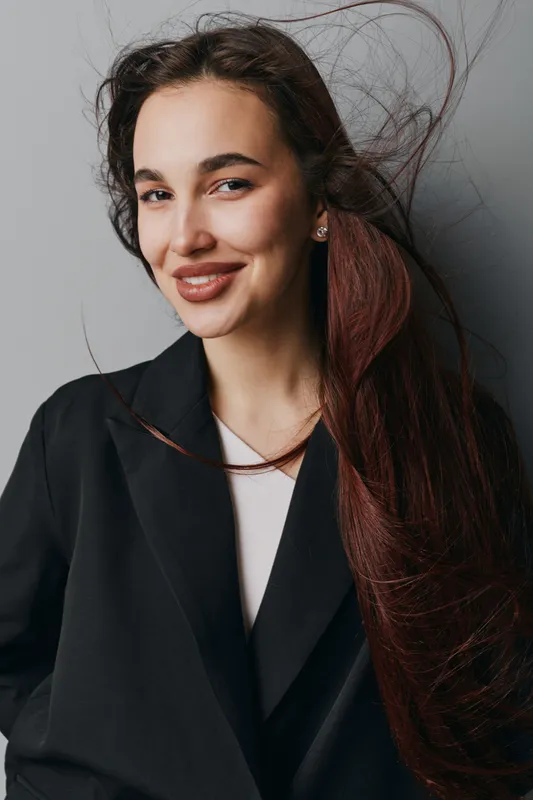 Woman with long brown hair wearing black blazer and white top smiling confidently in studio. Portrait of happy female model with natural makeup and earrings on gray background.