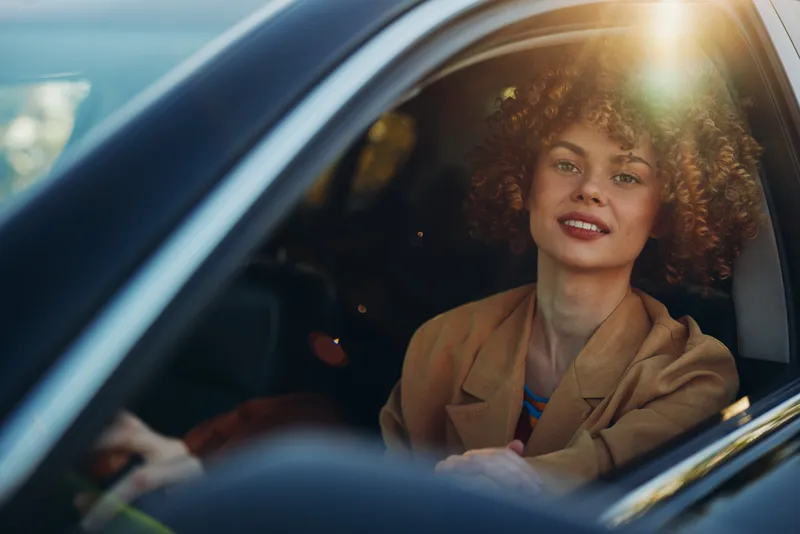 Smiling woman with curly hair sitting inside a car, driver poised in the front seat. Warm retro sunlight creates a nostalgic mood and relaxed lifestyle feeling.