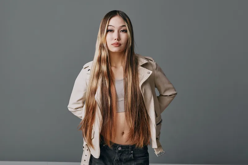 Young woman with long hair, wearing casual jacket and crop top, standing confidently against a plain background in studio. Fashion, beauty, and style portrait with natural expression.