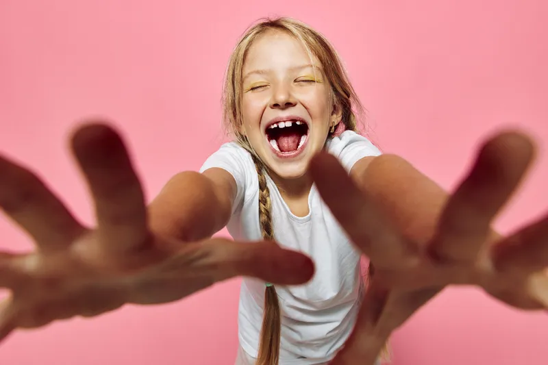 Happy girl with braided hair smiling widely with open arms against a pink background. Concept of joy, childhood, and playfulness.