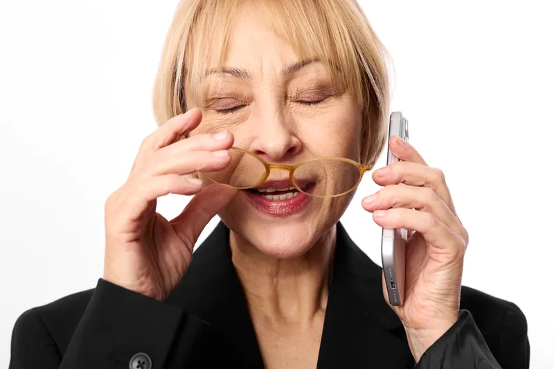 Stressed elderly woman in business suit speaks on mobile phone call, covers her nose with one hand, eyes closed and tired, concept of burnout and anxiety.