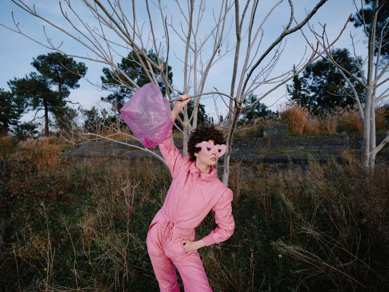 Pink suited dancer in an outdoor setting, windblown scarf and bold fashion meet artistic movement among bare trees and wild brush, conveying playful energy and surreal mood.