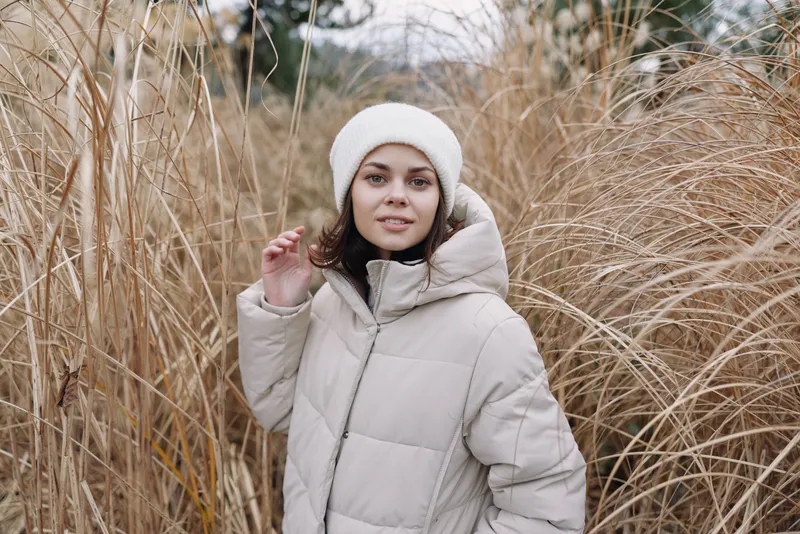 Winter outdoor portrait of a woman in a light puffer coat and white beanie among tall dried grasses, with soft morning light