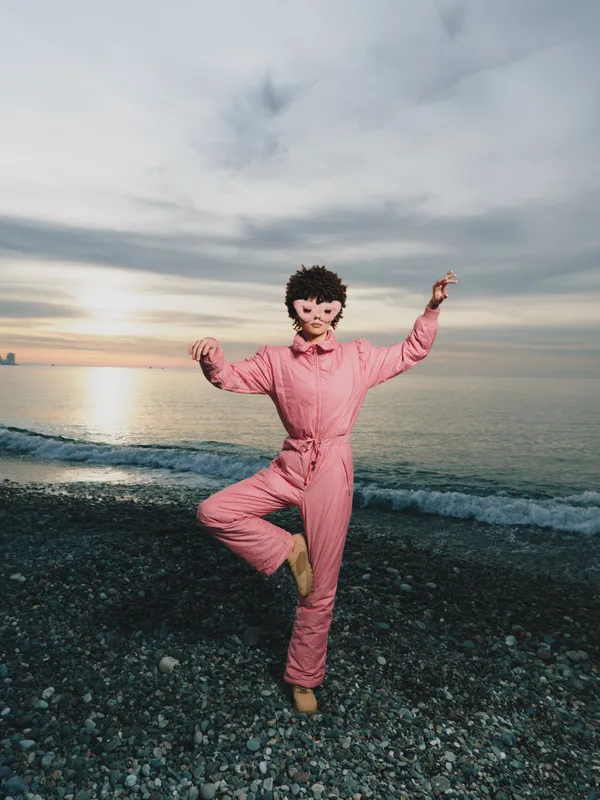 Person in pink jumpsuit dancing on a rocky beach at sunset, joyful pose with raised arms, carefree outdoor moment, sea horizon and soft light, vibrant lifestyle image for travel or wellness