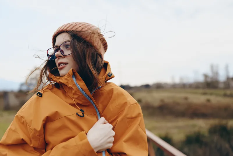 Woman lifestyle outdoors, wearing orange jacket and glasses, standing beside rural field and railway, wind lifts hair, calm portrait, soft daylight, natural colors, scenic horizon