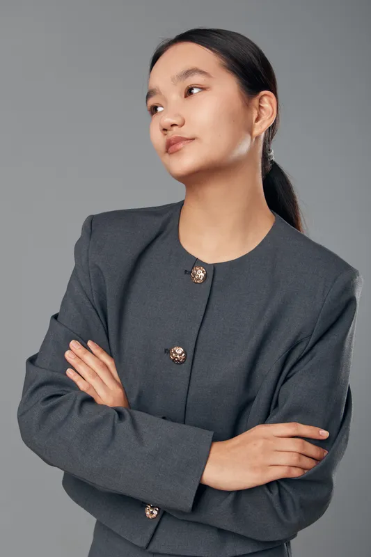Young woman with folded arms wearing gray business suit with buttons standing confident and thoughtful against gray background looking to the side. Professional female portrait.