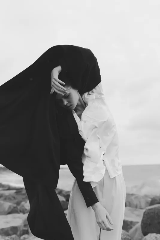 Veil and cloak drift over a figure by the shore, conveying mourning and mystery in a minimalist silhouette. White fabric contrasts with dark veil, capturing emotion, wind, solitude, and quiet drama