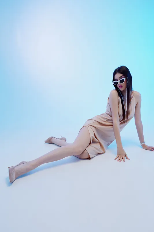 Young woman in a beige dress and sunglasses poses elegantly on the floor against a blue background. Concept of beauty, fashion, style, and modern trends in studio lighting.