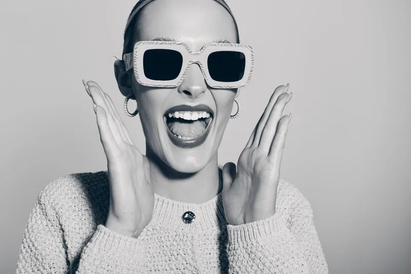 Happy white woman with pale skin wearing large sunglasses and cozy sweater expresses excitement with open mouth and raised hands on gray background.
