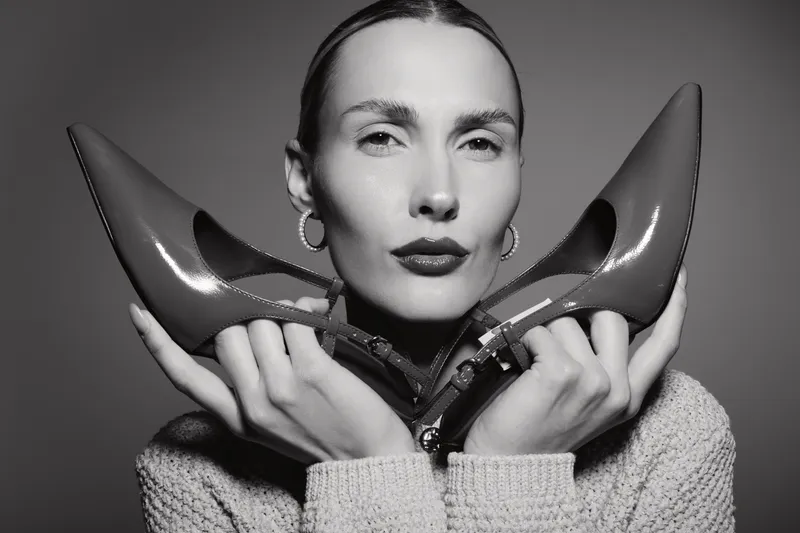 Portrait of white woman with pale skin holding pair of pointed shoes near her face in black and white studio image.