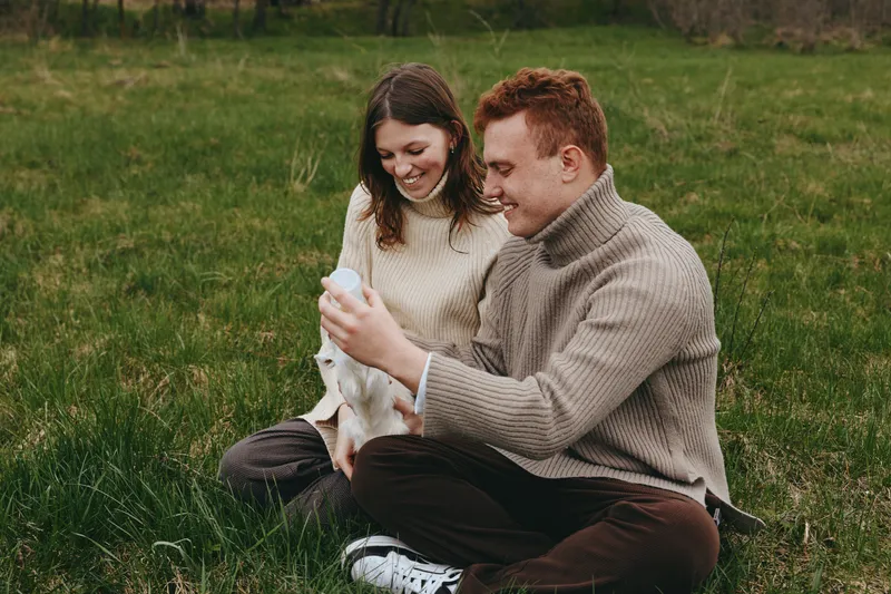 A happy couple sits on grass in a sunlit field, smiling at a small goat kid between them. A warm, candid moment reflecting countryside lifestyle, nature, and gentle bonding with animals.