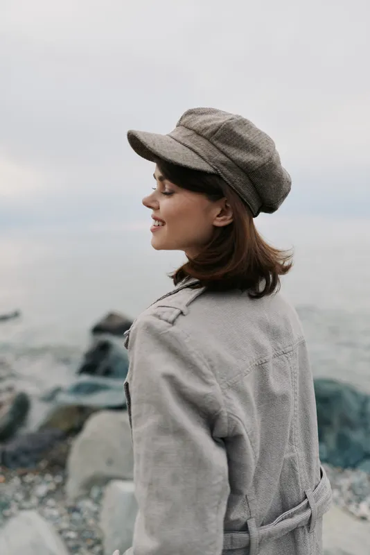 Young woman in a cozy coat and hat stands near rocky shore looking sideways, smiling gently. Overcast sky and calm sea background create peaceful outdoor atmosphere.