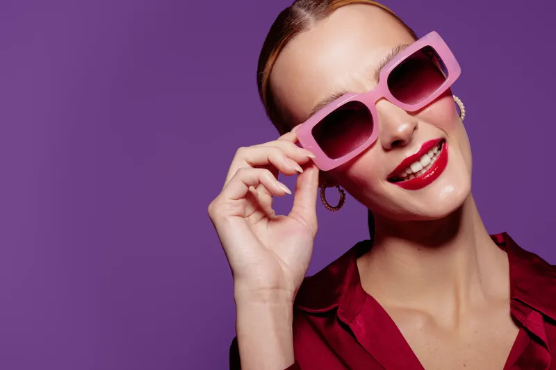 Stylish woman with red lipstick wearing pink sunglasses and red blouse smiles confidently against purple background. Concept of fashion, beauty, summer style, and accessories.