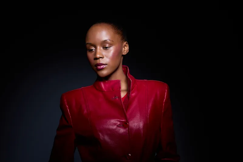 Portrait of a confident dark skinned African woman wearing a red leather jacket against a black background. The studio lighting highlights her smooth skin and stylish outfit.